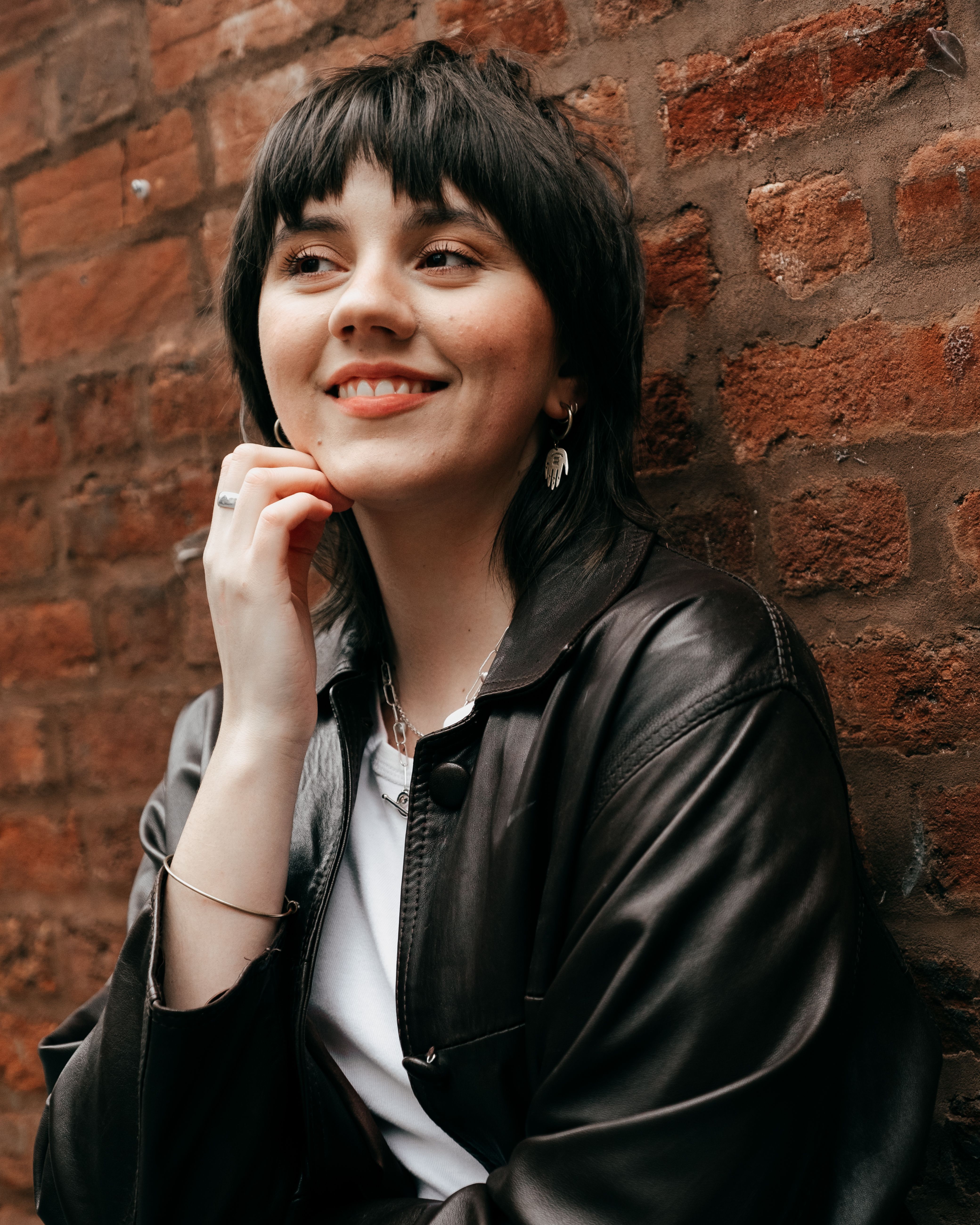 Charming young female with short dark hair in trendy leather jacket touching face and looking away against brick wall in daylight