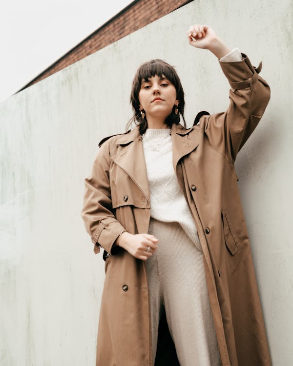 Free From below of confident young woman with short dark hair in stylish trench coat standing with raised arm against concrete wall in daylight Stock Photo