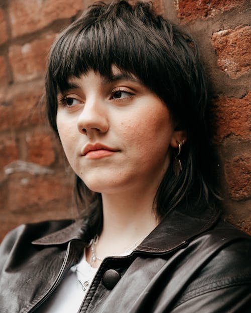 Thoughtful young female in leather jacket looking away against brick wall