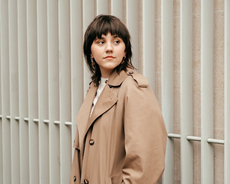 Calm Young Woman In Trench Coat Looking Away Against Wall In Daytime