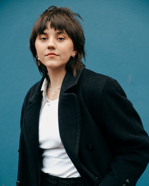 Confident female with hairstyle and trendy clothes standing against blue wall of building and looking at camera in daytime