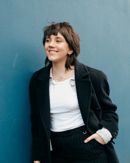 Positive woman with hairstyle in trendy clothes standing with hand in pocket and leaning on blue wall in daytime