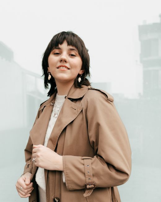 Self assured young female millennial with dark hair in trendy trench coat smiling and looking at camera while standing on street near building with glass walls