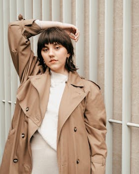 Young woman in a trench coat posing against a fence, urban and serene mood.