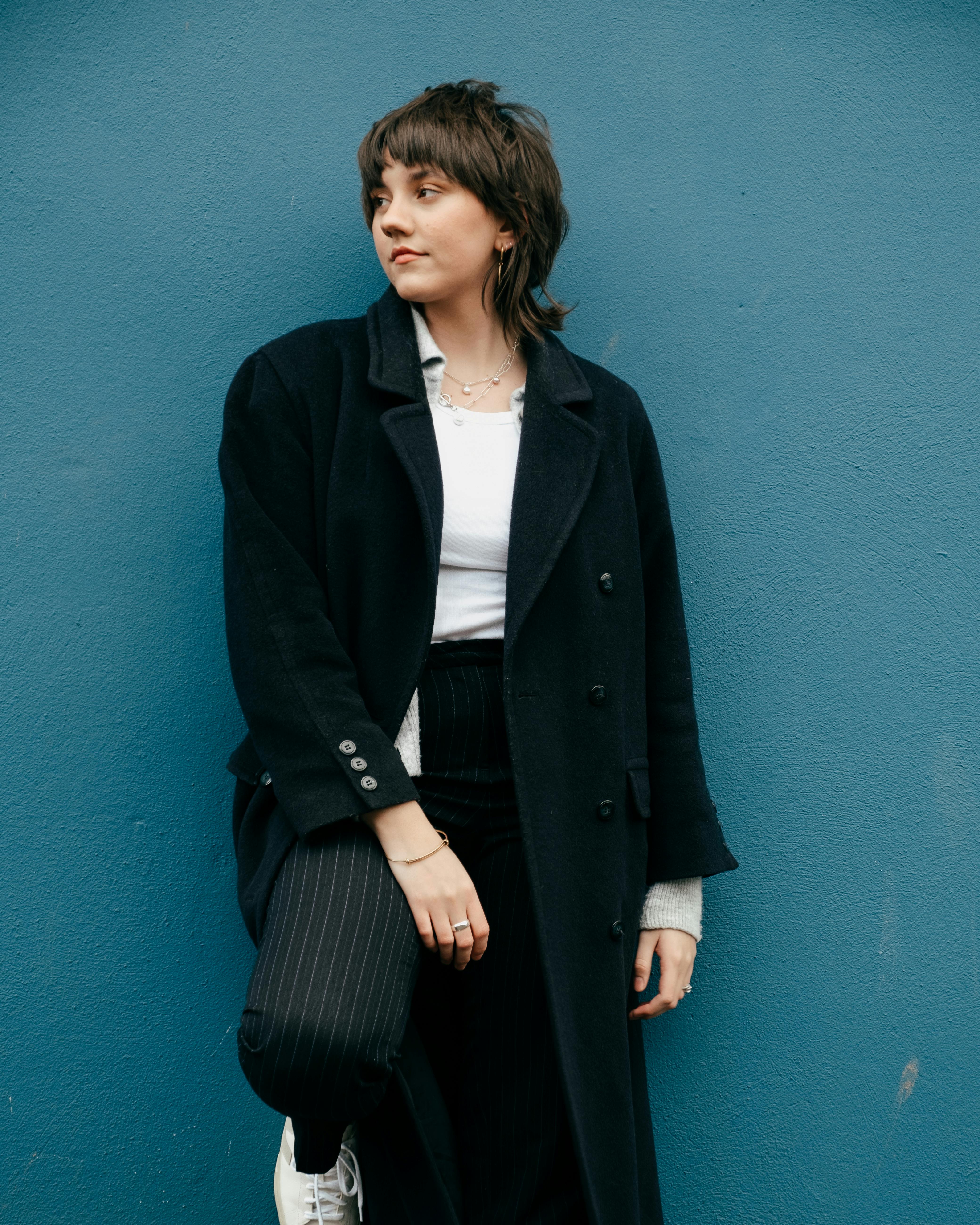 Stylish young female in trendy clothes and coat looking away while standing near stone blue wall in street in daylight