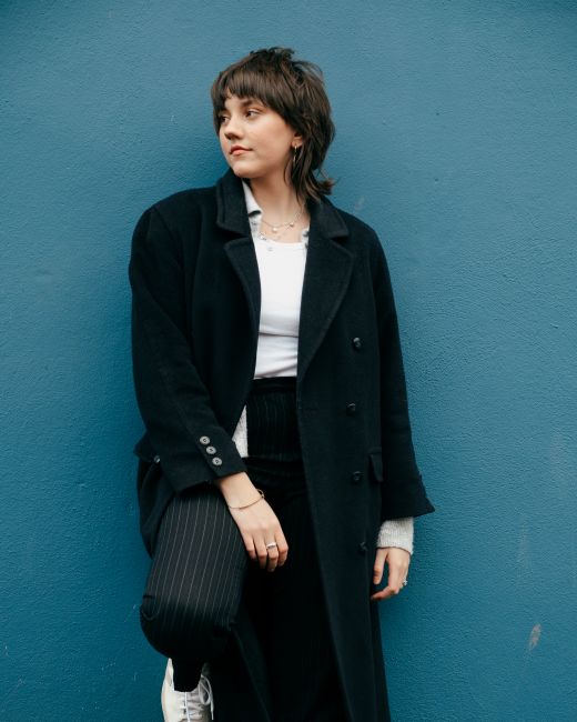 Stylish young female in trendy clothes and coat looking away while standing near stone blue wall in street in daylight
