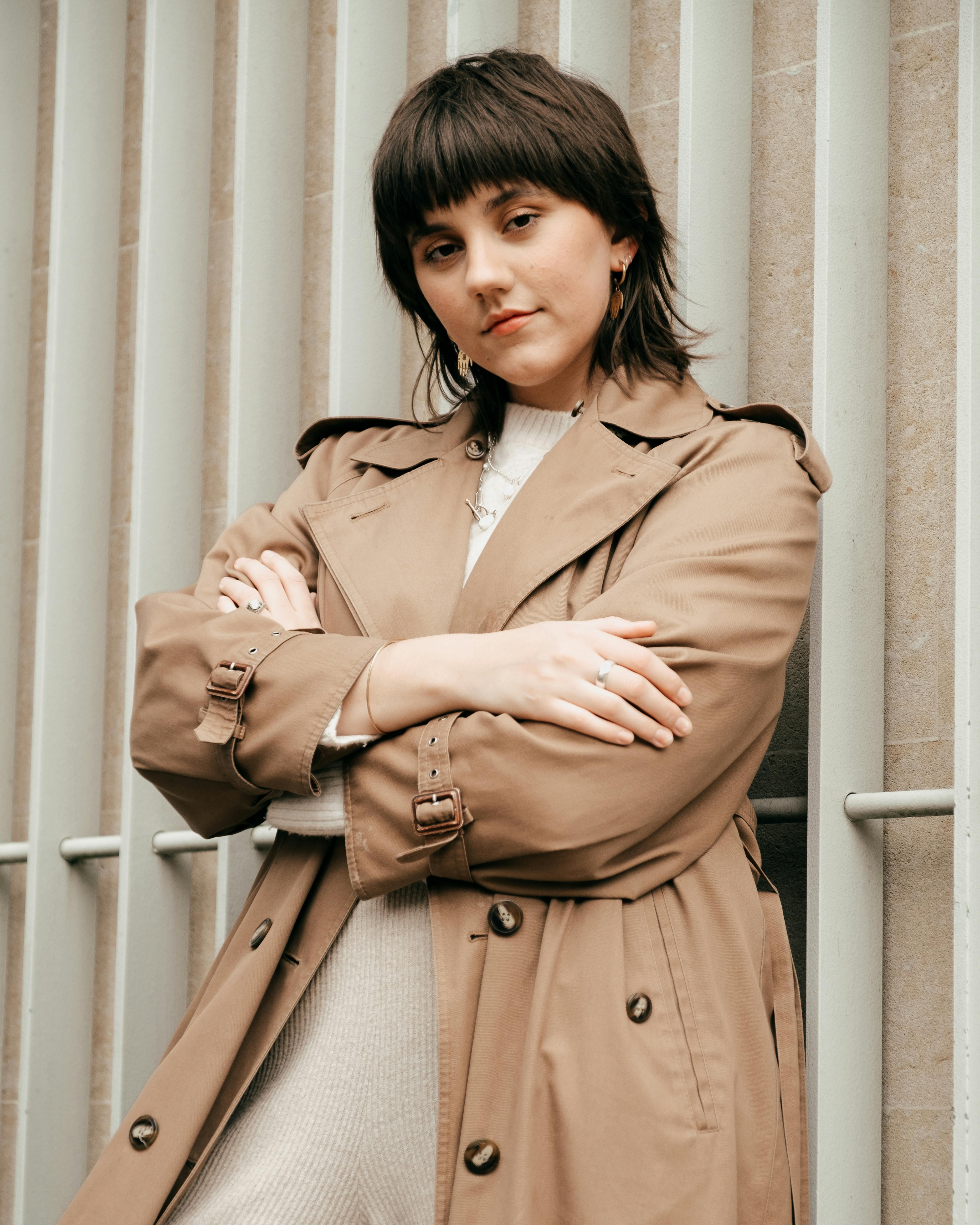 Confident young woman wearing trendy coat while standing near metal lattice and wall with crossed hands in street in daytime while looking at camera