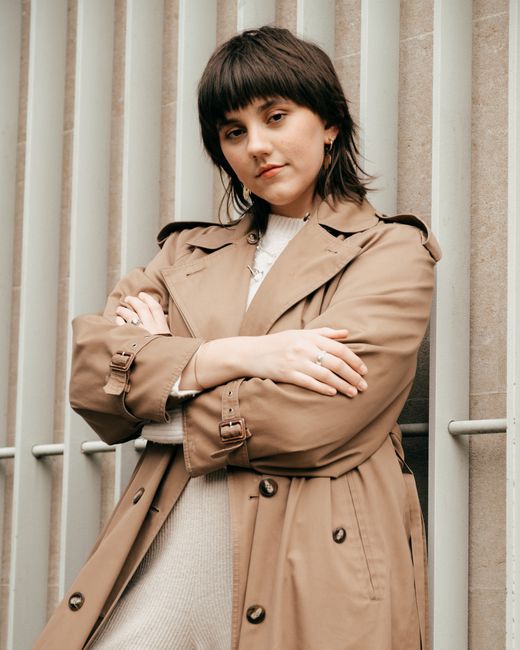 Confident young woman wearing trendy coat while standing near metal lattice and wall with crossed hands in street in daytime while looking at camera