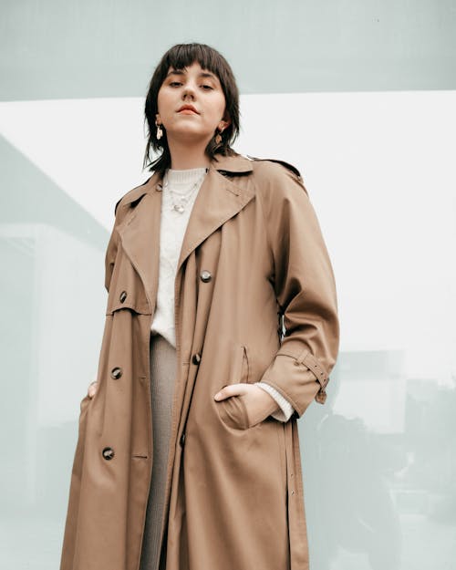 Low angle of confident stylish young female with dark hair in trendy coat standing on street with hands in pockets and looking at camera