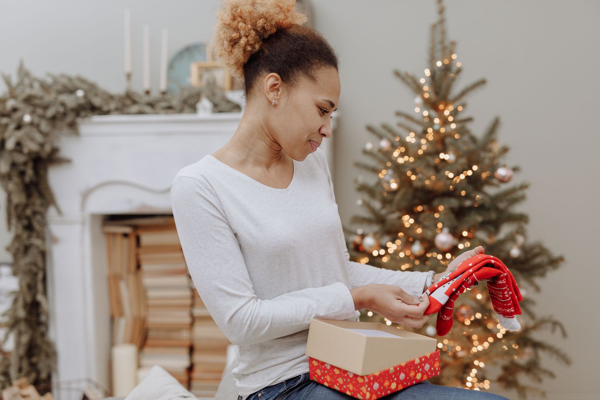 Smiling woman sits by a fireplace with a gift box and a Christmas tree decorated in the background.