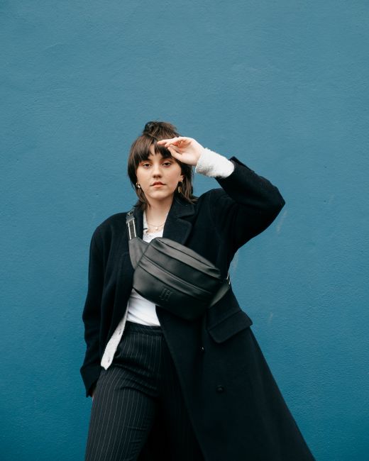 Confident young woman wearing trendy coat and waist bag while standing near blue stone wall with raised hand while looking at camera in street in daytime