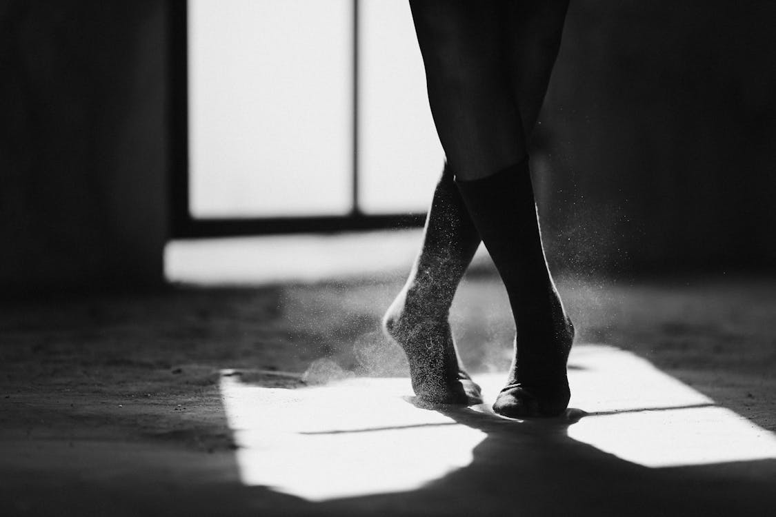 Free Black and white of crop anonymous female ballet dancer standing on dusty floor with crossed legs Stock Photo