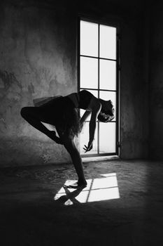 Black and white side view of flexible female ballet artist performing choreographic movement while leaning back with raised leg in building