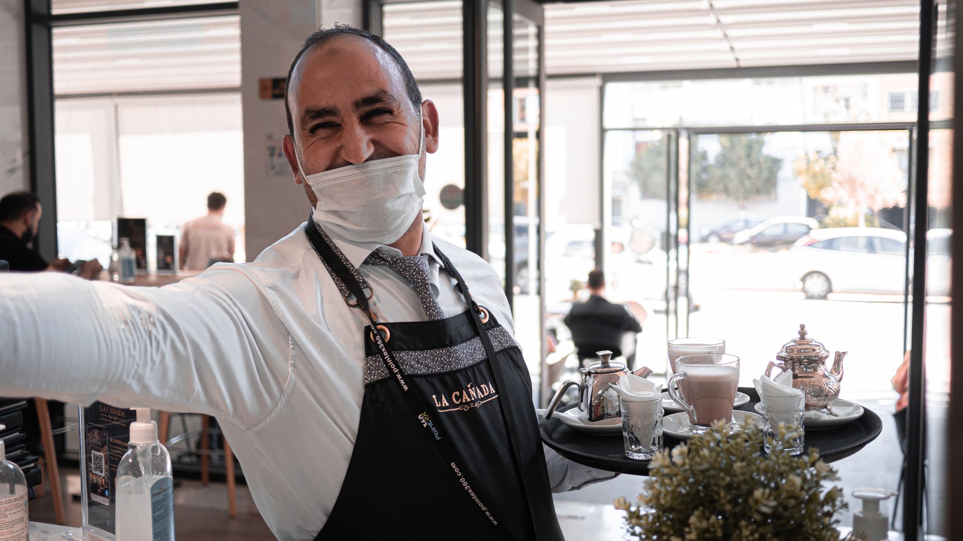 A cheerful waiter with a face mask serves drinks on a tray in a cafe in Fes, Morocco.