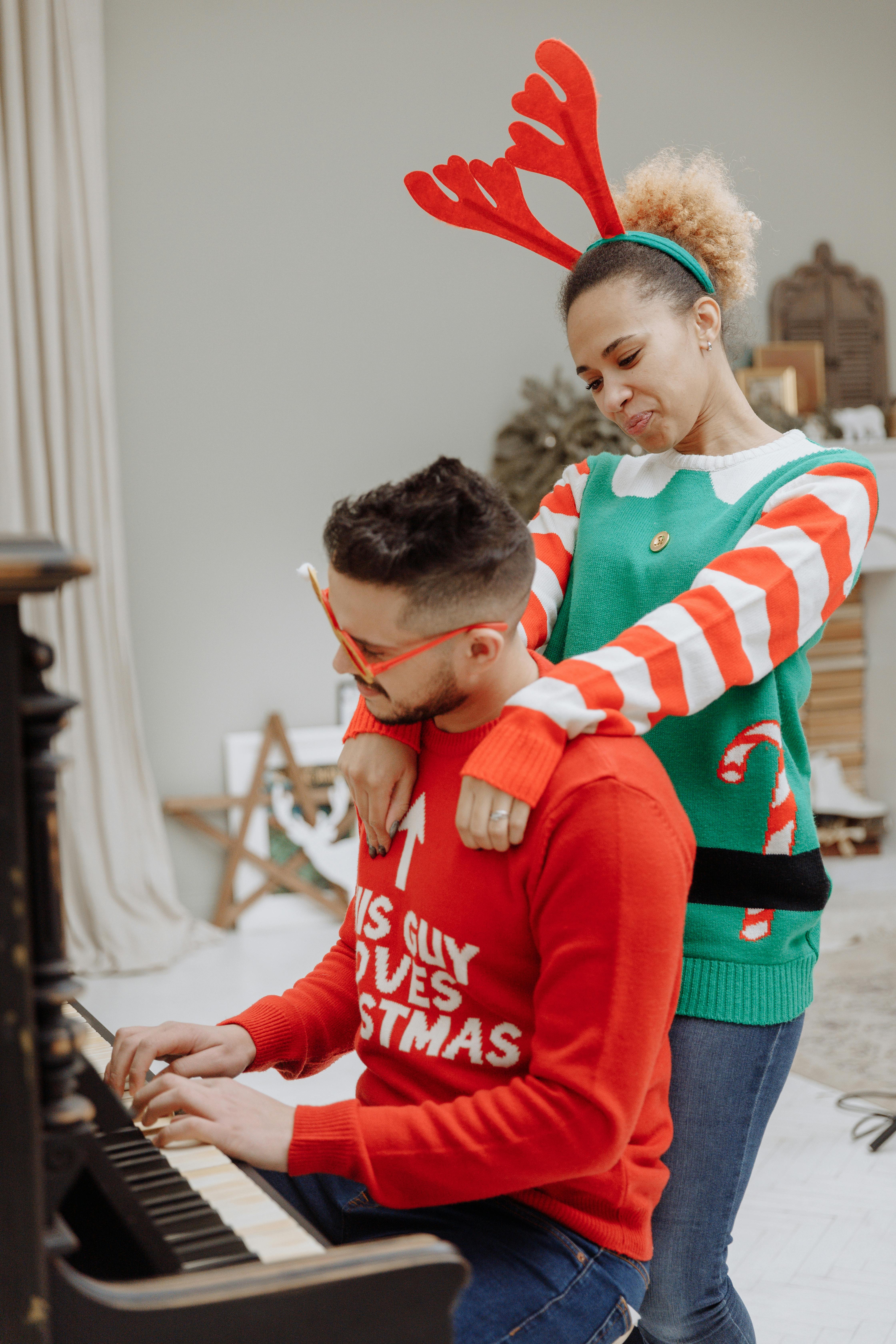 Couple wearing festive sweaters enjoying music by the piano indoors during the holiday season.