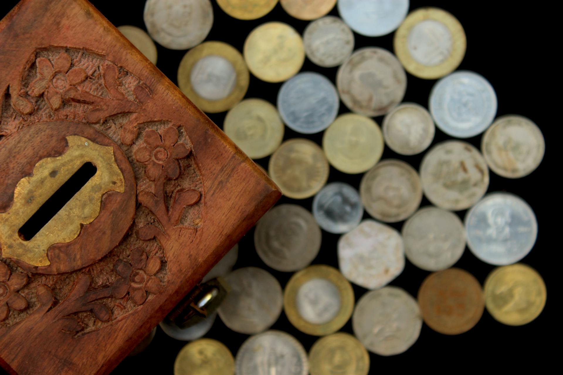 Top view of a carved wooden money box surrounded by assorted coins on a dark background.