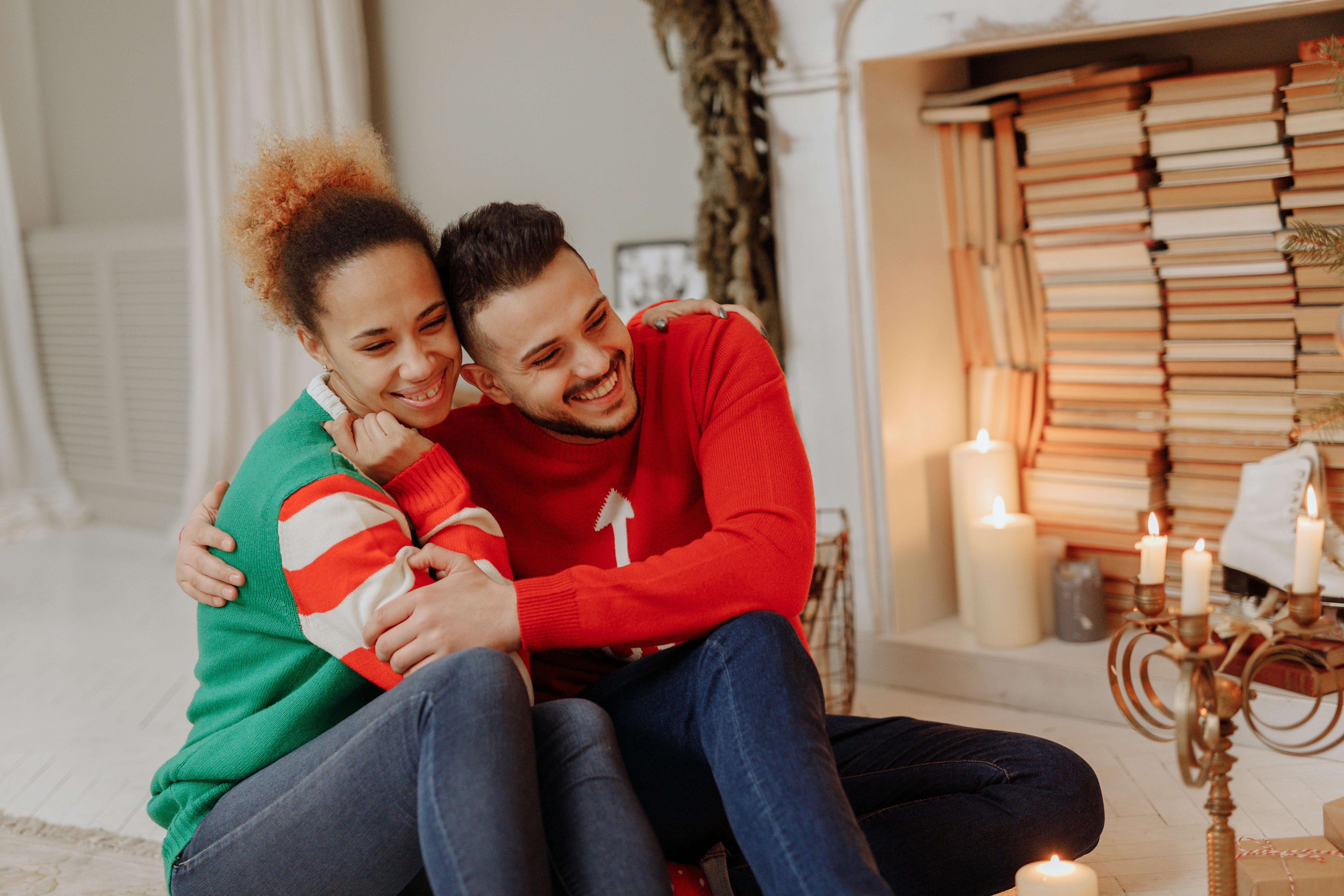 Joyful couple in cozy sweaters embracing in a warm, festive setting by the fireplace.