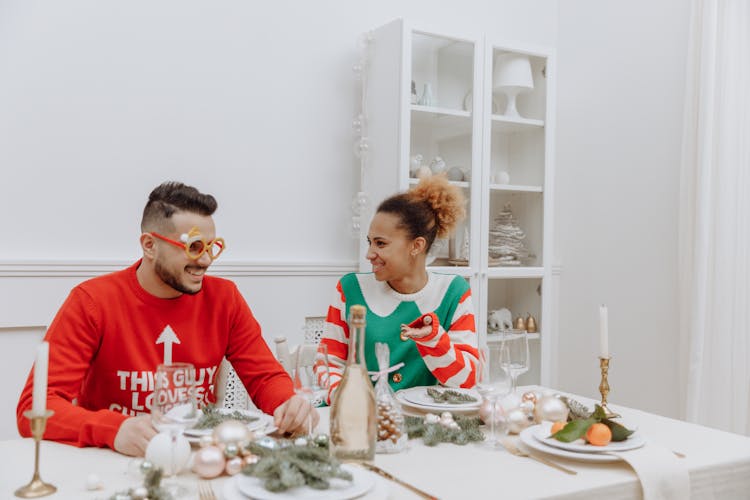 A Couple In Ugly Christmas Sweaters Sitting At The Table