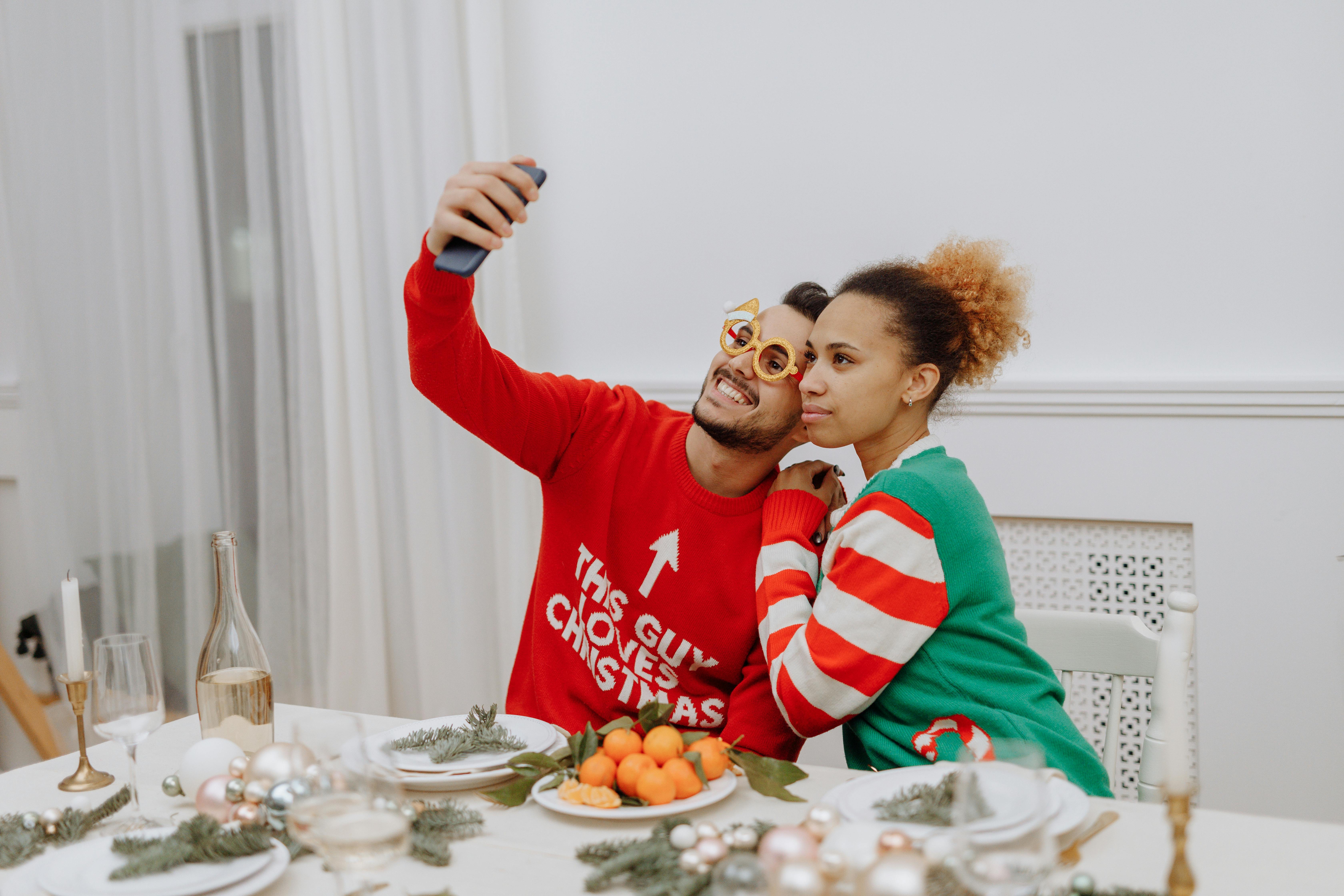 Smiling couple taking a selfie in festive Christmas sweaters during a holiday gathering indoors.