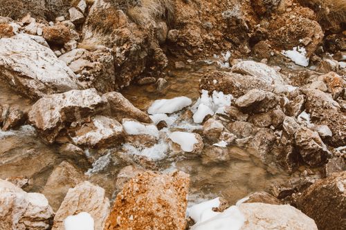 Free A picturesque winter stream flows over snowy and rocky terrain, showing nature's raw beauty. Stock Photo