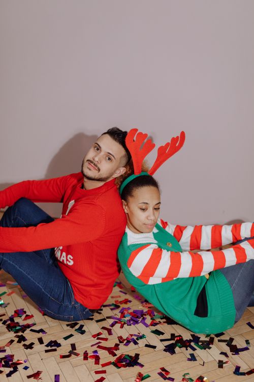 Free A cheerful couple in Christmas sweaters seated on a confetti-covered floor, celebrating holidays Stock Photo