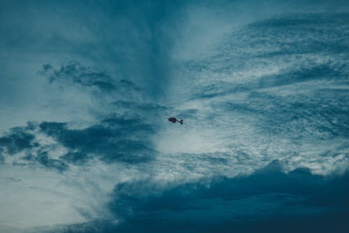 From below silhouette of distant helicopter with propeller flying in gloomy sky with floating fluffy clouds at sunset time in nature