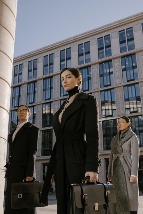 Free Three professional women in coats carrying briefcases stand confidently in front of a modern office building. Stock Photo