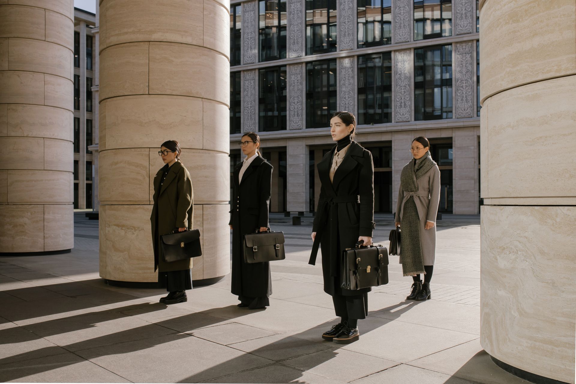 Group of businesswomen in formal attire with briefcases, standing outdoors near columns.