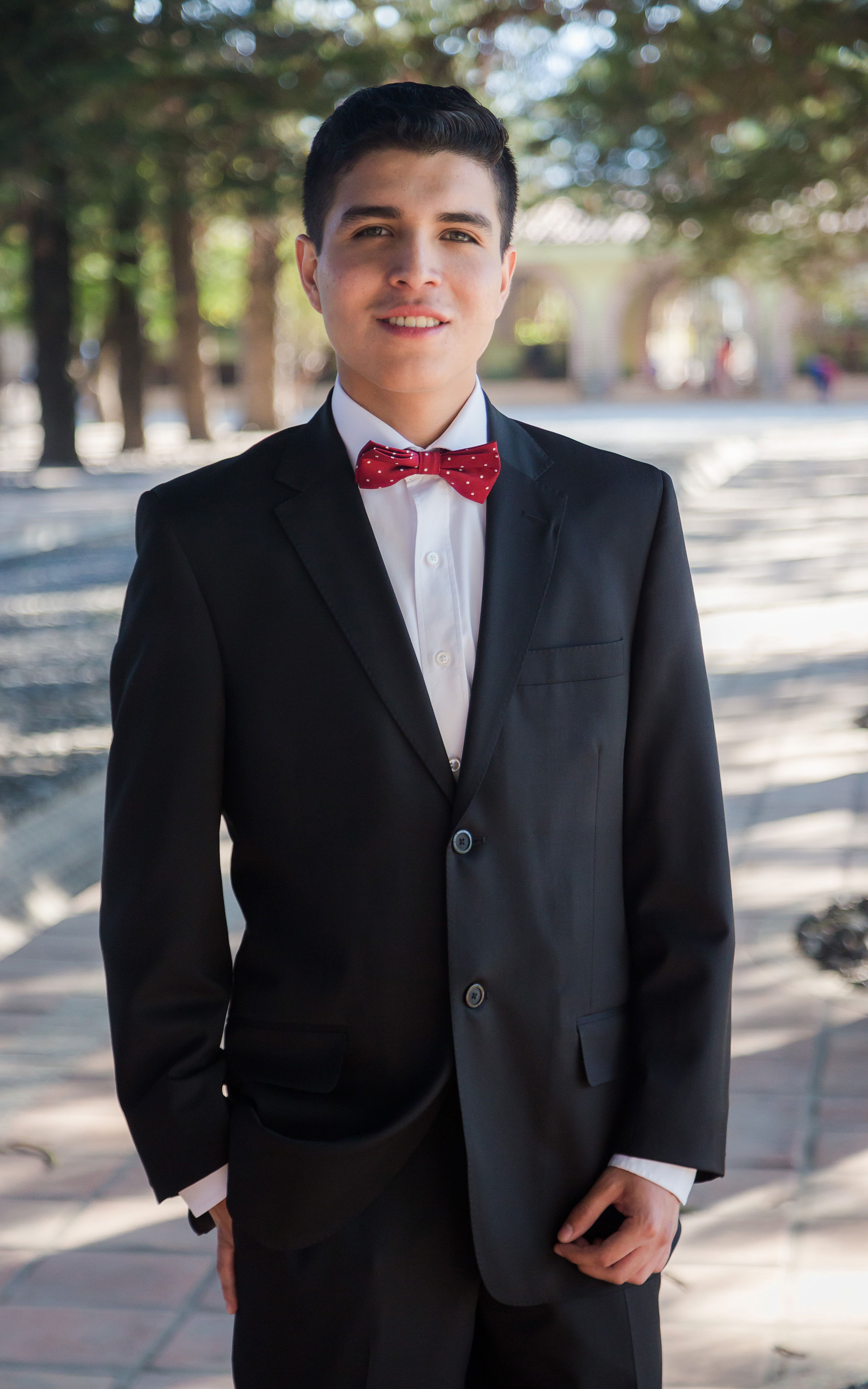 Young man in black suit with red bow tie smiling outdoors.