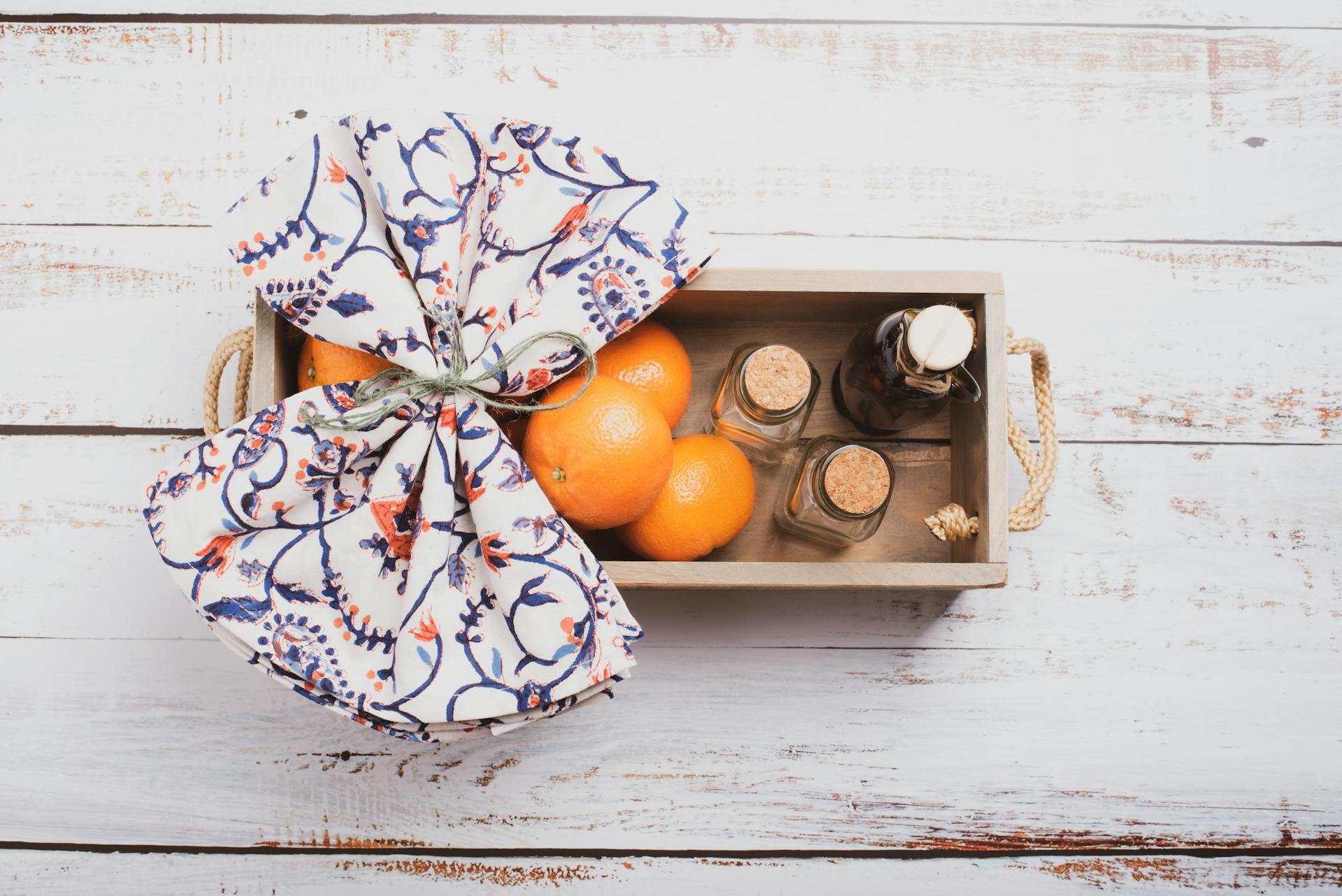 A rustic wooden box filled with fresh oranges and decorative bottles, wrapped in a patterned cloth.