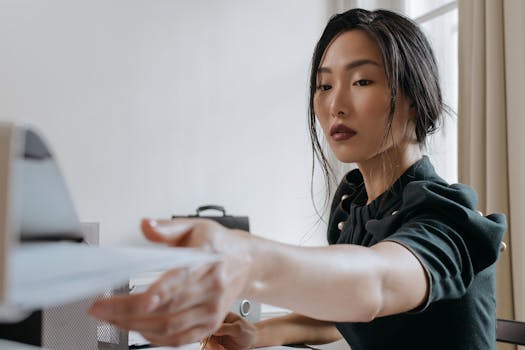 Asian business woman reaching for document in office setting, demonstrating focus and professionalism.
