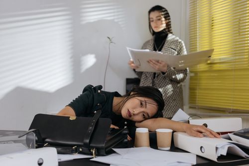 Free Exhausted employee resting on desk surrounded by documents, colleague in background. Stock Photo