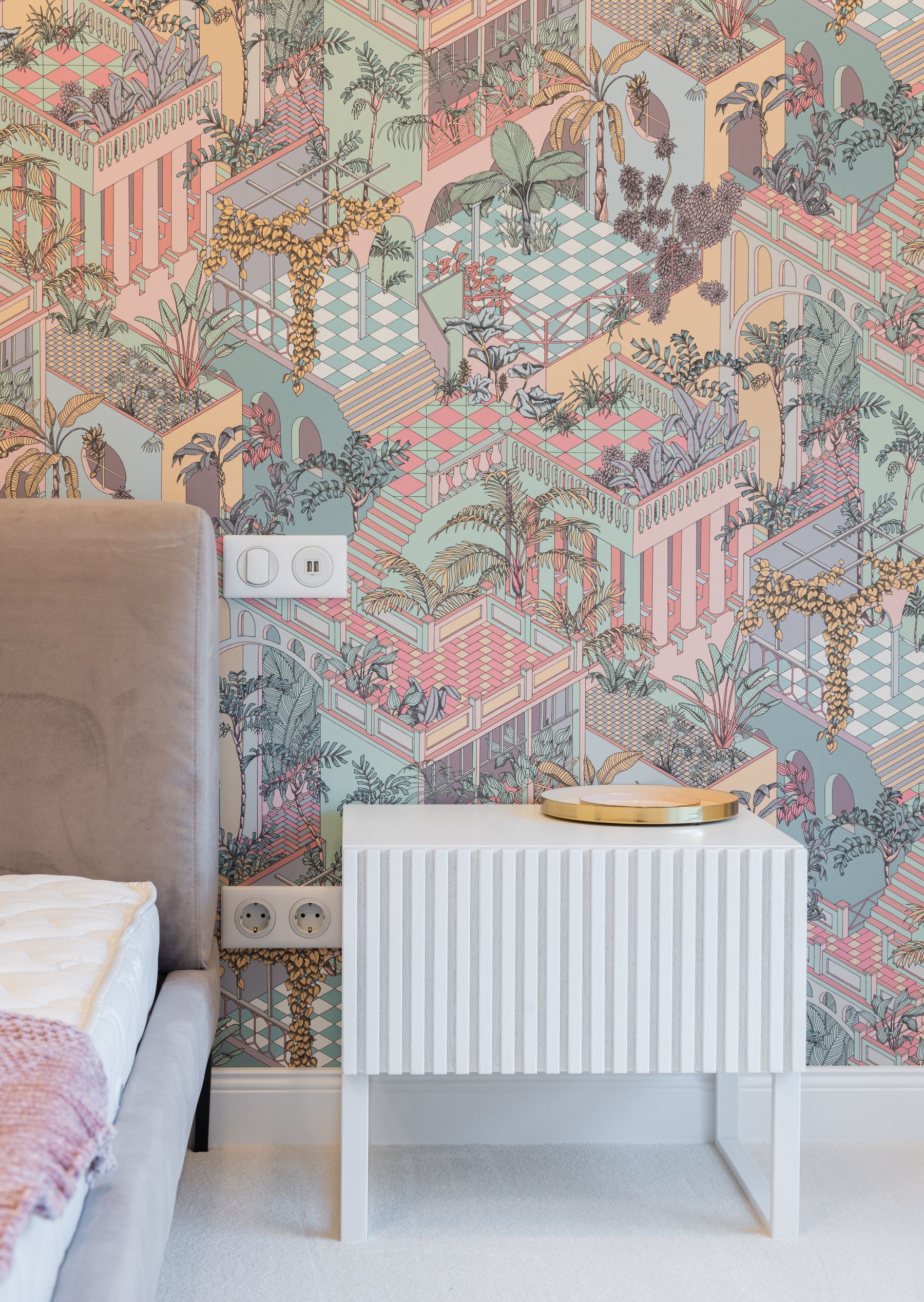 Compact bedroom detail with multicolor patterned wallpaper, pale upholstered bed edge and white radiator cover