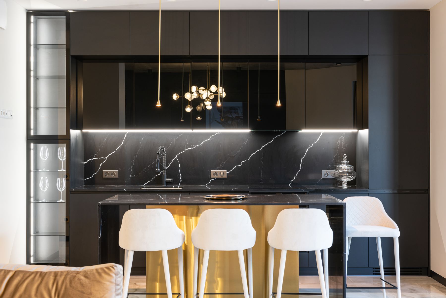 Dark kitchen cabinets and black veined backsplash behind white bar chairs and a lit timber-toned island face