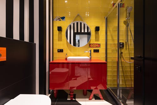 Interior of modern bathroom with sink on red cabinet near tap and mirror hanging on yellow wall near toilet and shower