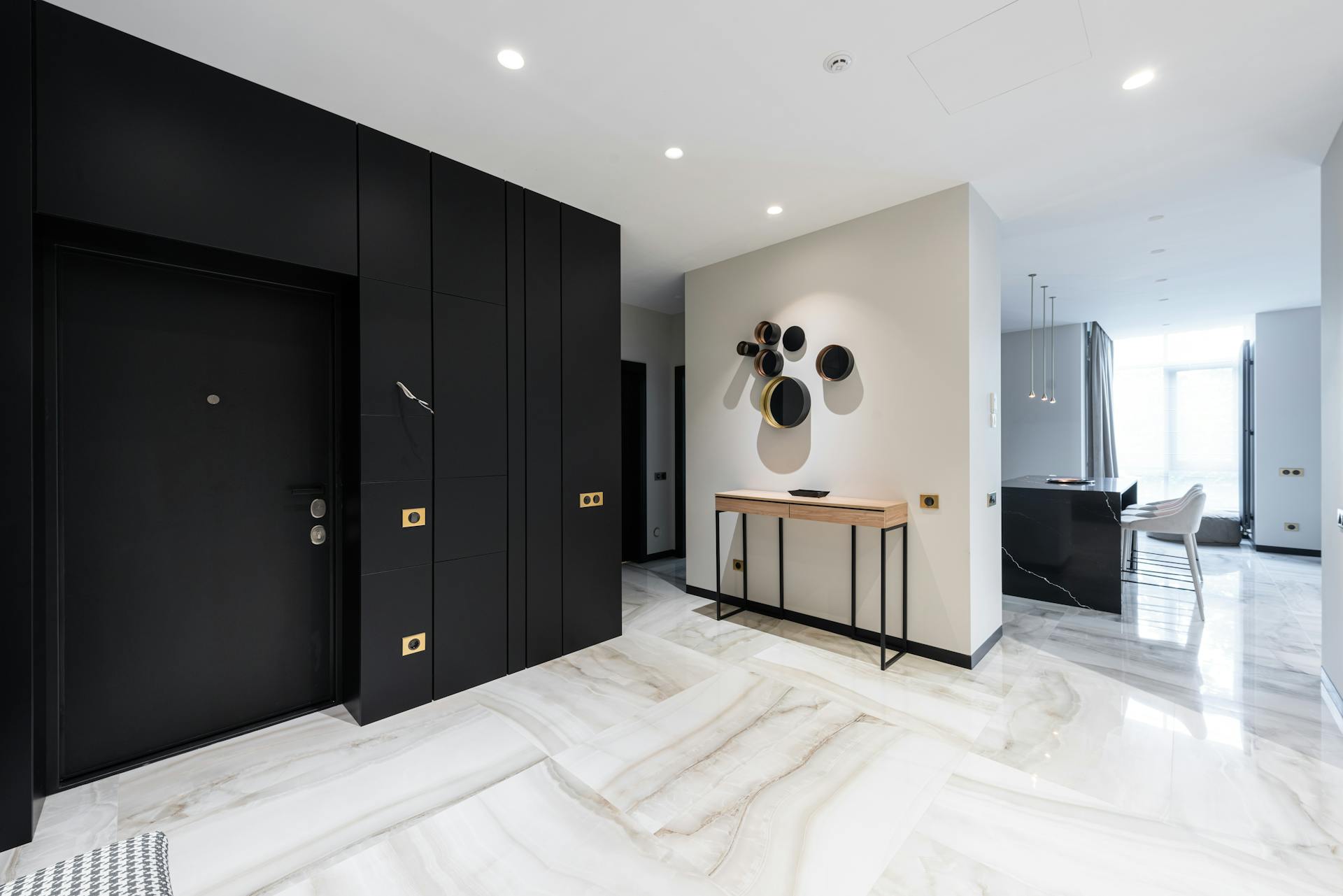 Interior of modern light hall with black doorway and stylish table at wall with entrance to open plan kitchen with black island conuter