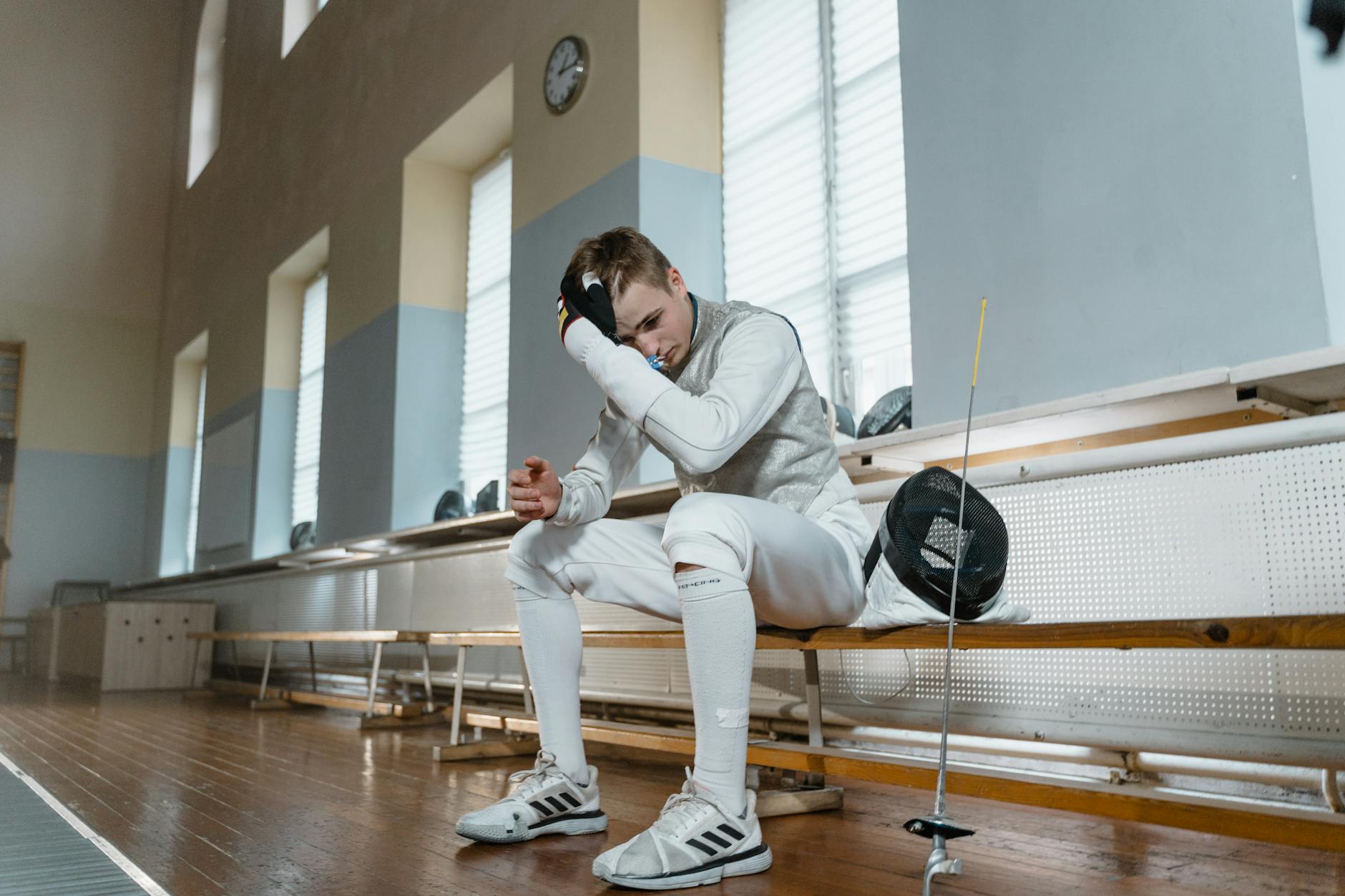 Young male fencer taking a break in a training salle, wearing full fencing gear.