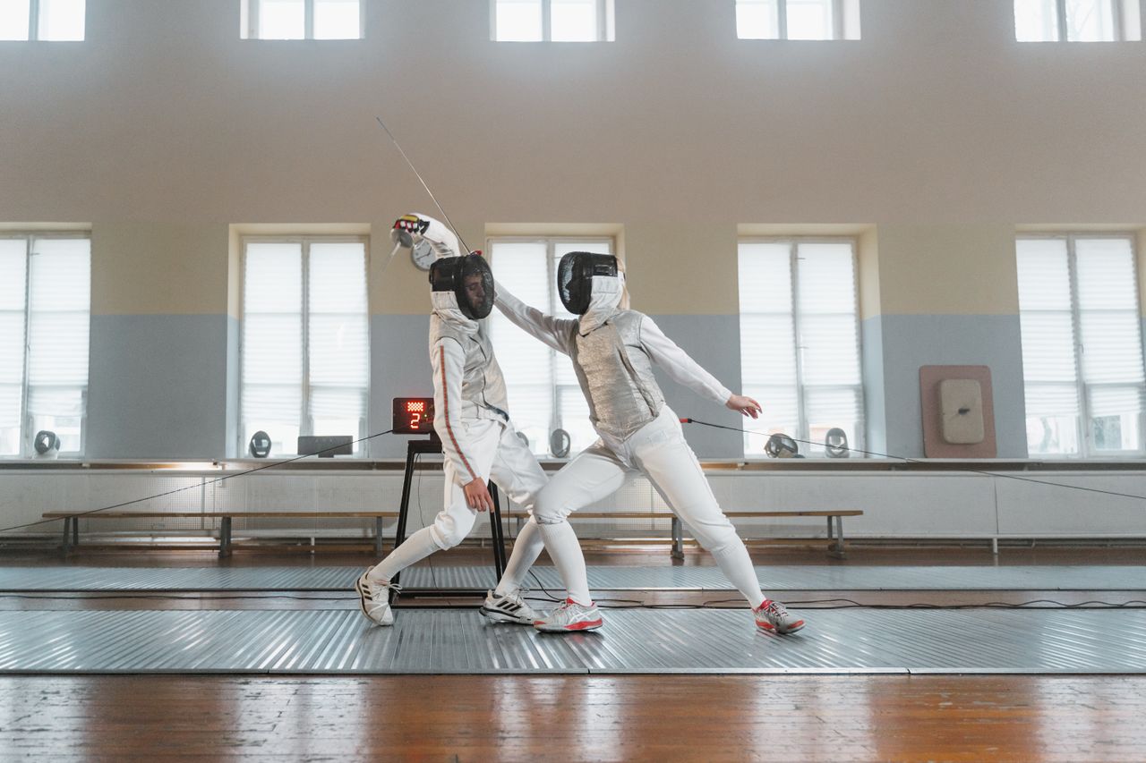 two fencers engage in a duel indoors with bright natural lighting
