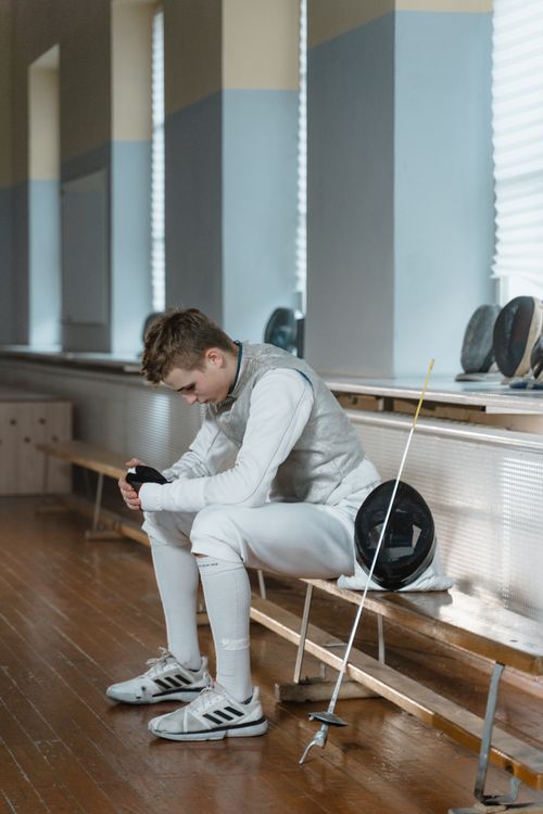 Free A fencer in full gear resting on a bench indoors with fencing equipment by their side. Stock Photo