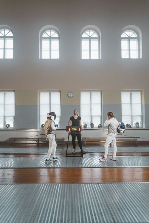 Free Two women fencing in an indoor sports facility monitored by a referee. Stock Photo