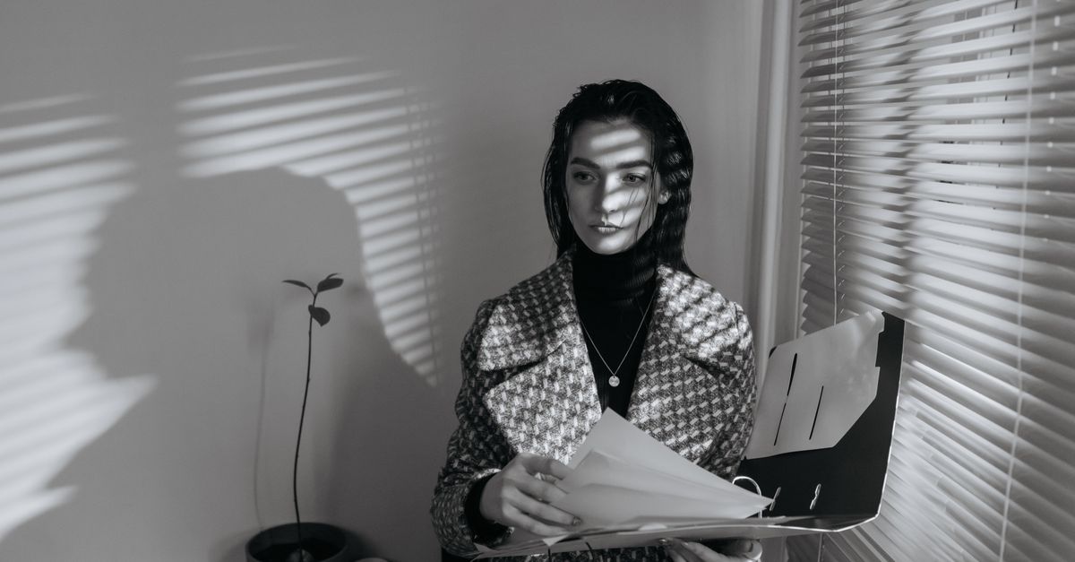 Mujer de negocios en blanco y negro, revisando documentos con persianas que crean sombras.