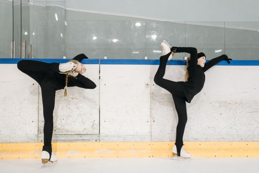 Two young female figure skaters practicing stretches on an indoor ice rink.