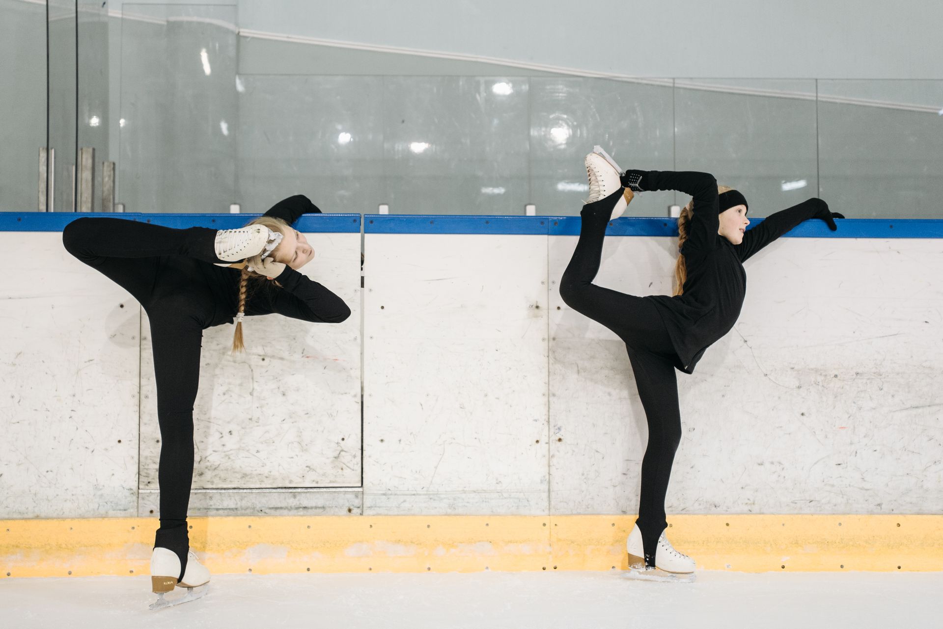 Two young female figure skaters practicing stretches on an indoor ice rink.