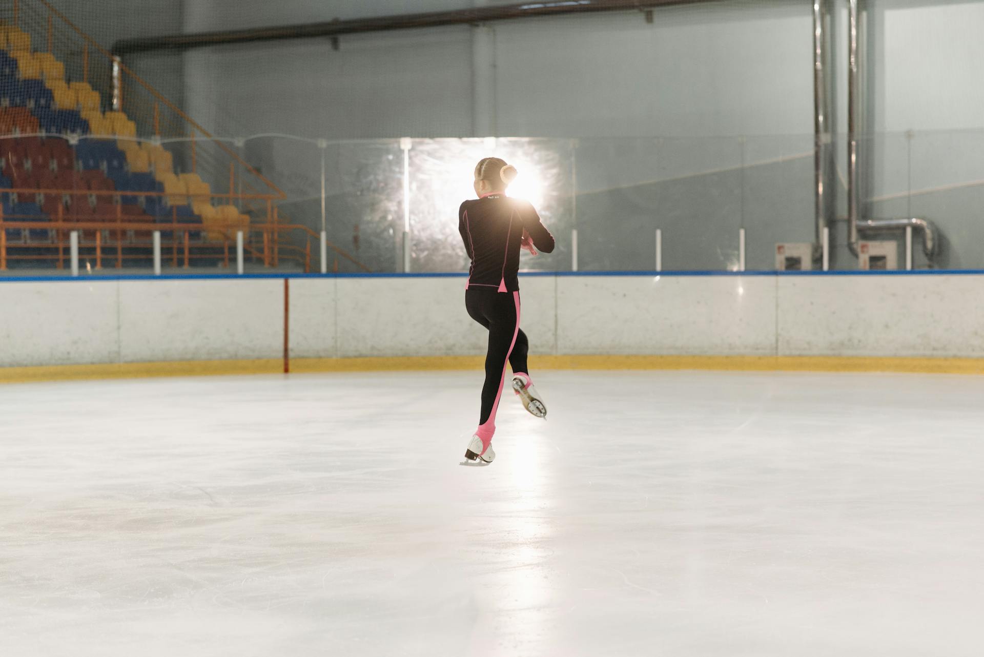 A female figure skater gracefully performs a jump on an indoor ice rink, backlit by arena lights.