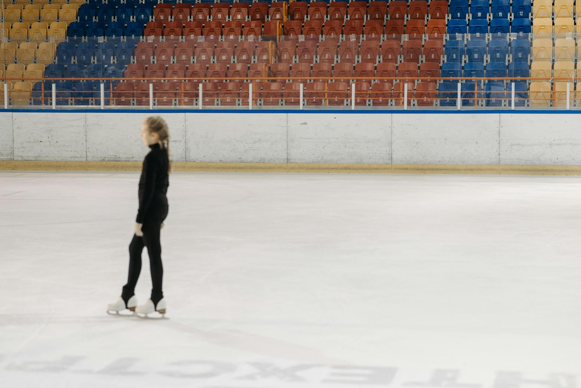 A young figure skater practicing on an indoor ice rink with colorful seating in the background.