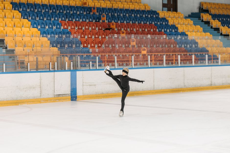 A child figure skater executes a skillful move on an empty ice rink with colorful seating.