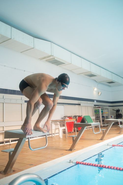 Free A male swimmer in goggles and cap prepares to dive from a block at an indoor swimming pool. Stock Photo