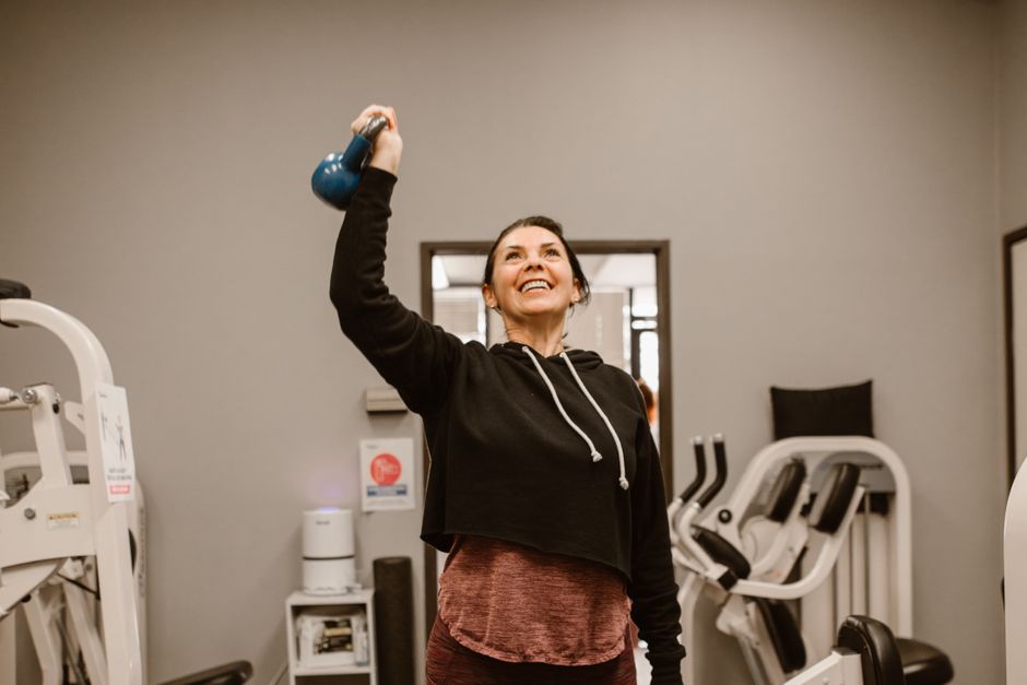 Smiling woman lifting a kettlebell in a gym setting, showcasing fitness and well-being.