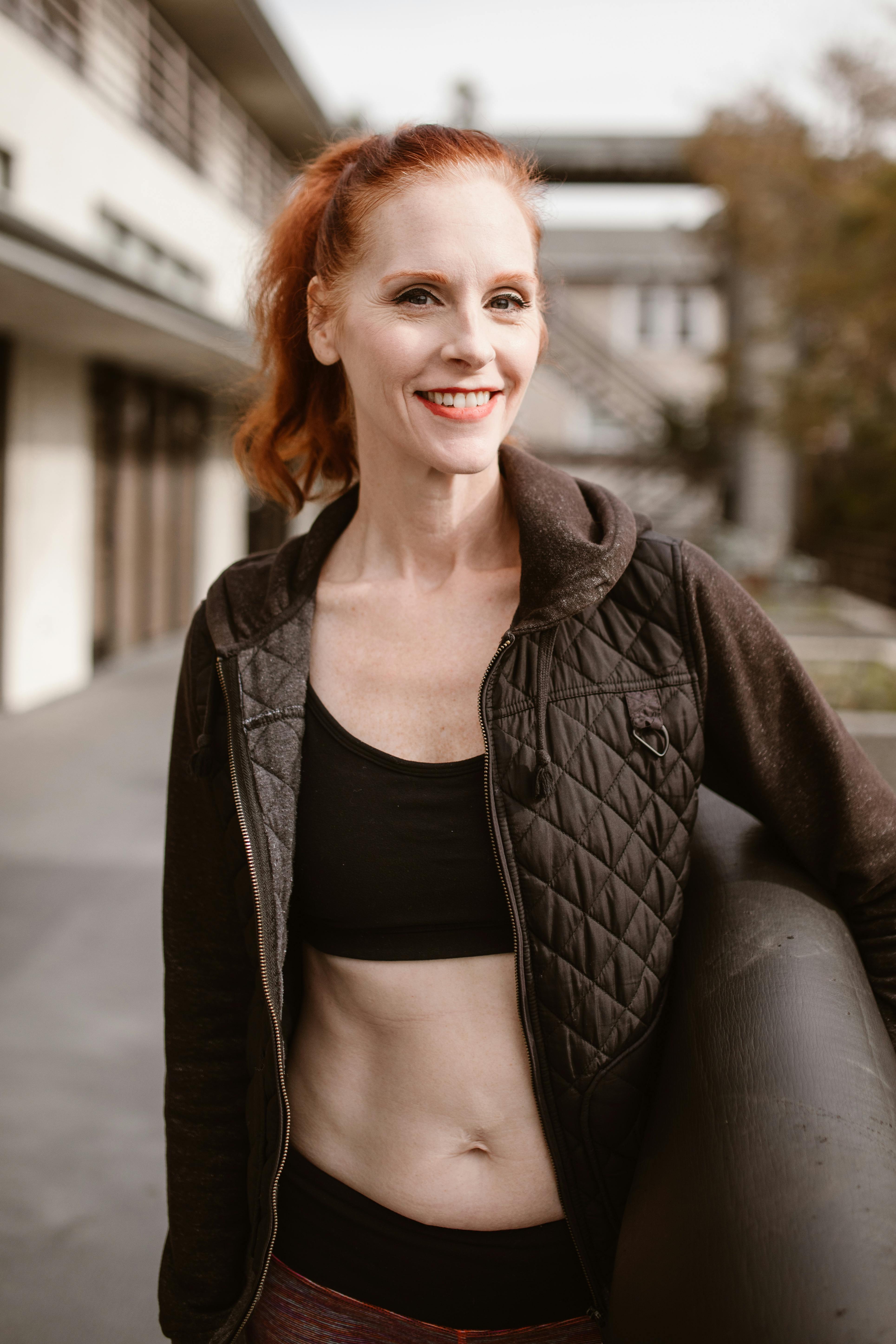 Woman in a black jacket and activewear smiling while holding a yoga mat outdoors.