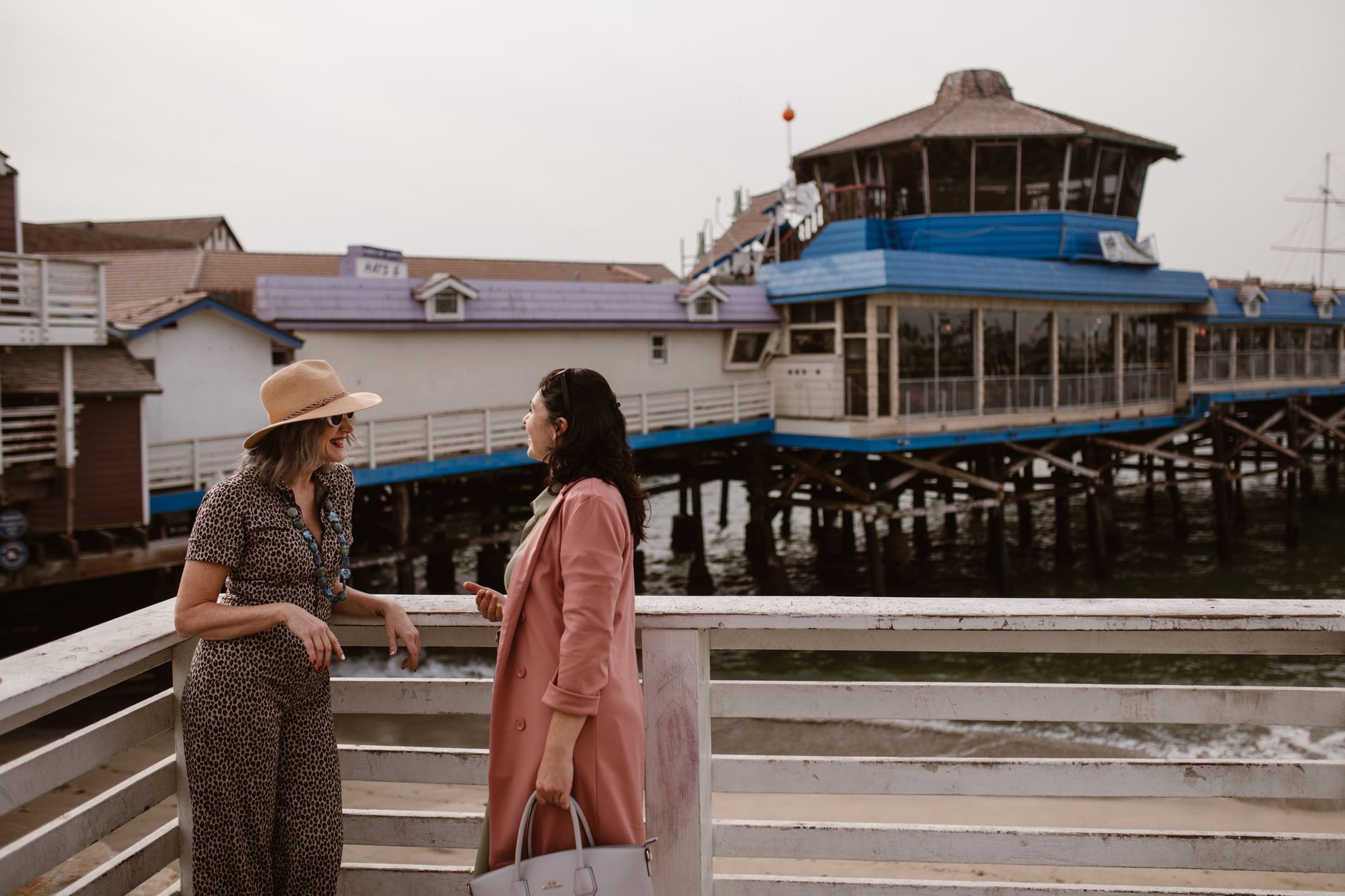 Two women enjoy a lively conversation on a wooden pier by a waterfront restaurant.